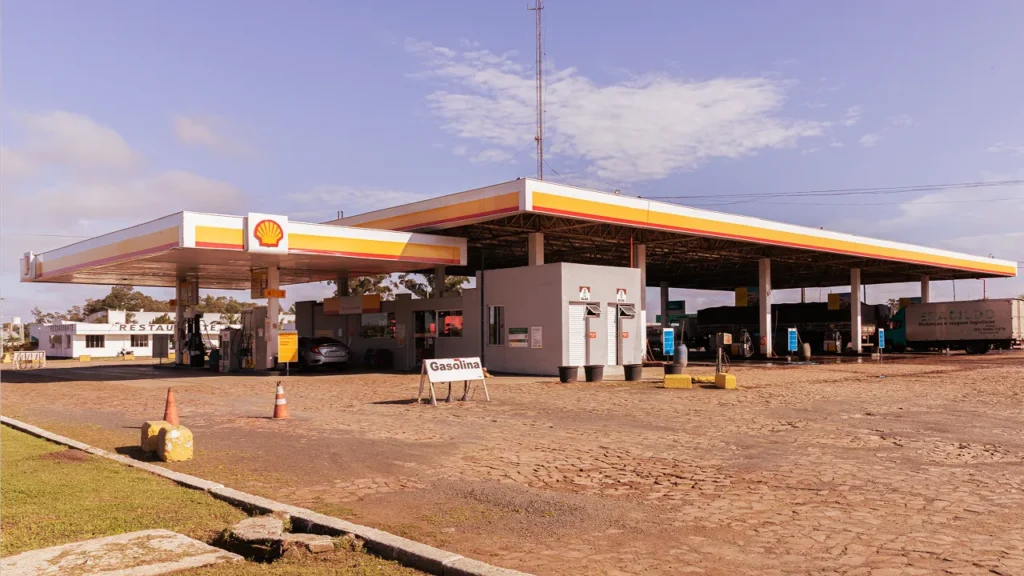 Vista principal do posto de gasolina Shell com grandes coberturas, bombas e sinalização de Gasolina.