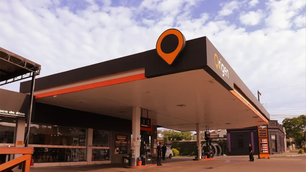 Vista principal da fachada do posto Origen, com a grande placa preta e o pino de localização laranja no topo, e a área de abastecimento.